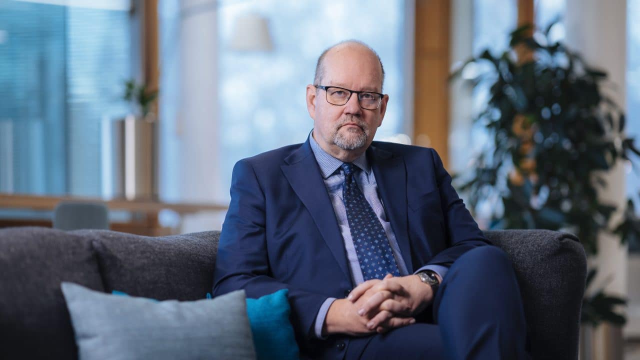 Kuvassa parrakas mies, jolla on silmälasit ja siniset puvuntakki, paita ja solmio, istuu sohvalla. Jaakko Kiander