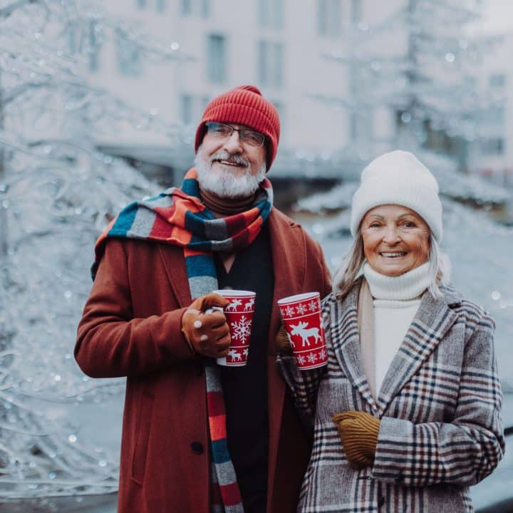Kuvassa hymyilevä vanhempi pariskunta värikkäissä talvivaatteissa juomamukit kädessä.